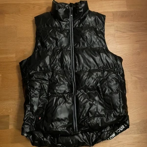 Svart dunväst med dragkedja - Snygg svart dunväst med dragkedja framtill. Västen har en hög krage och praktiska fickor. Perfekt för kyligare dagar när du vill hålla stilen.