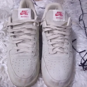 Beige Nike Air Force 1 - Säljer ett par klassiska Nike Air Force 1 i beige färg. Skorna har en stilren design med snörning och den ikoniska Nike-loggan på sidan. Perfekta för en avslappnad stil.