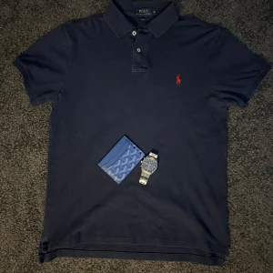 Mörkblå pikétröja från Ralph Lauren - Säljer en stilren mörkblå pikétröja från Ralph Lauren med klassisk krage och knappar. Tröjan har en röd broderad logotyp på bröstet och är i slim fit. Perfekt för en avslappnad men ändå uppklädd look. Storlek M och bra skick!