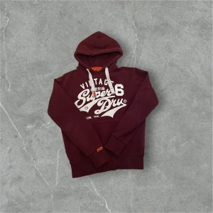 Vinröd hoodie från Superdry - Säljer en vinröd hoodie från Superdry med vit text och logga på framsidan. Storlek: S Skick:9/10