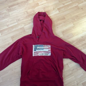 Röd hoodie från Ralph Lauren - skön röd hoodie från Ralph Lauren med en stor logga på bröstet som har ett amerikanskt flaggmotiv med slitningar och text. Väldigt svår och få tag på. Nypris ligger runt 2500 Mitt pris 699. Priset är inte hugget i stenen vid snabb affär
