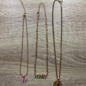 Tre guldhalsband med olika hängen - Säljer tre stiliga guldhalsband. Ett med en rosa fjäril, ett med texten 'HONEY' och ett med ett runt medaljonghänge. Perfekta för att ge din outfit en personlig touch.