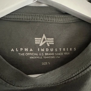 Grå t-shirt från Alpha Industries - Säljer en stilren grå t-shirt från Alpha Industries i storlek S. T-shirten har en klassisk passform med korta ärmar och en liten logga på bröstet. Perfekt för en avslappnad look.