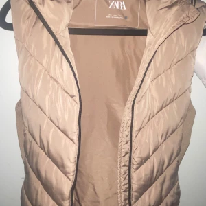 Beige dunväst från Zara - Snygg beige dunväst från Zara med dragkedja framtill. Perfekt för lager-på-lager under kyligare dagar. Västen har en stilren design och är lätt att matcha med olika outfits.