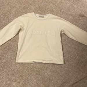 Vit långärmad tröja från Moncler - Säljer en stilren vit långärmad tröja från Moncler med deras logga i glittriga bokstäver på framsidan. Perfekt för en casual look med en touch av lyx. Tröjan har en rund halsringning och är tillverkad i ett mjukt material.