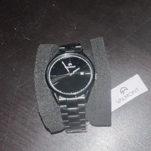 Svart klocka från Valmont - Snygg ,stilren och vattentät svart klocka från Valmont med datumvisning.  Klockan har en svart urtavla och ett matchande armband i rostfritt stål. Klockan är alldrig använd. Perfekt för den som gillar en klassisk look med modern touch. Utgångspris är 3119kr, jag säljer varan för 2600