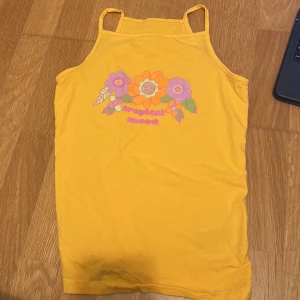 Gul linne med blommigt tryck - Säljer en solgul linne med smala axelband och ett färgglatt blommigt tryck framtill. Perfekt för sommardagar med sin lätta och bekväma design. Texten 'tropical mood' ger en härlig sommarkänsla.