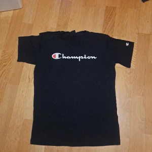Mörkblå t-shirt från Champion - Säljer en svart t-shirt från Champion med deras ikoniska logga i vitt och rött på bröstet. T-shirten har korta ärmar och är perfekt för en avslappnad stil. Klassisk design som funkar till det mesta.