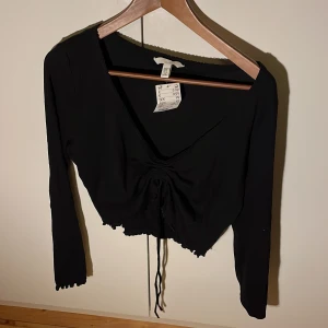 Svart långärmad topp från H&M - Säljer en svart långärmad topp från H&M med v-ringning och knytning framtill. Toppen har en croppad design och är perfekt för en stilren look. Passar bra till både jeans och kjol.