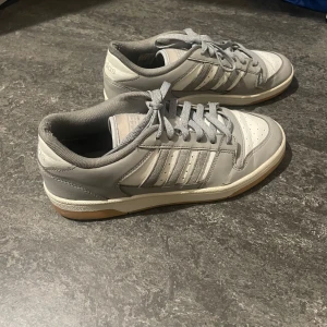 Grå och vita sneakers från Adidas - Snygga grå och vita sneakers från Adidas med klassiska ränder på sidan. Skorna har en gummisula i beige och snörning framtill. Perfekta för en avslappnad stil.