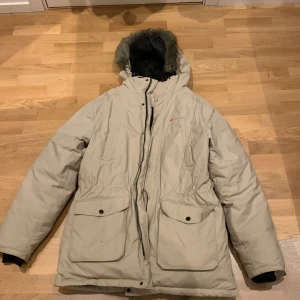 Beige parkajacka med pälsdetalj - Säljer en beige parkajacka med huva och pälsdetalj. Jackan har dragkedja och knappar framtill samt stora fickor med lock. Perfekt för kalla dagar. Priset kan diskuteras