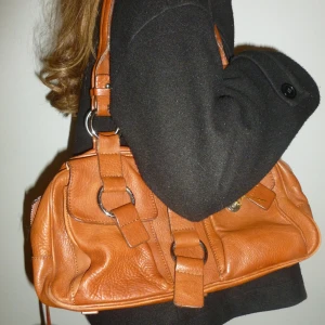 Brun skinnväska med detaljer - Snygg brun skinnväska med dubbla handtag och stiliga metallringar. Väskan har flera spännen och en praktisk dragkedja för säker förvaring. Perfekt för att ge din outfit en chic touch.