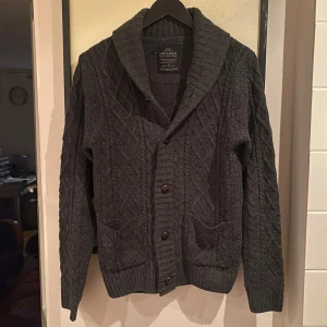 French Cable Cardigan - Säljer en snygg mörkgrå kabelstickad cardigan från Threadbare Denim i färgen mörkgrå. Cardiganen har två praktiska fickor framtill. Perfekt för vardagen eller festliga tillfällen! Storlek S och i dunder skick!
