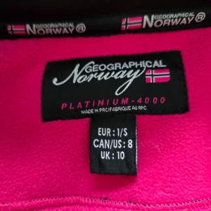 Svart jacka från Geographical Norway - Säljer en svart jacka från Geographical Norway med rosa detaljer och dragkedjor. Jackan har en huva och flera praktiska fickor. Perfekt för kyliga dagar med sin Turbo-Dry teknologi.