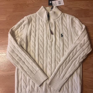 Vit stickad tröja från Ralph Lauren - Säljer en stilren vit stickad tröja från Ralph Lauren med kabelstickat mönster och dragkedja vid halsen. Tröjan har långa ärmar och en broderad logotyp på bröstet. Perfekt för kyliga dagar!(Priset är inte förhandlingsbart. Prutare ignoreras! 700 som gäller.)