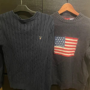 Mörkblå tröjor från Gant och Ralph Lauren - Två mörkblå tröjor, båda är från Gant och den ena har kabelstickat mönster och broderad logga och har storleken S, o den andra har en amerikansk flagga på bröstet och är i storleken M. Båda har långa ärmar och rund halsringning. Köp en för 400 eller båda för 599.