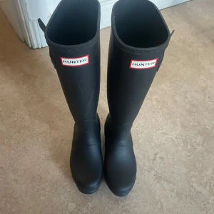 Svarta stövlar från Hunter - Snygga svarta stövlar från Hunter med klassisk design. Perfekta för regniga dagar med hög skaft och justerbar rem på sidan. Hunter-loggan syns tydligt framtill.