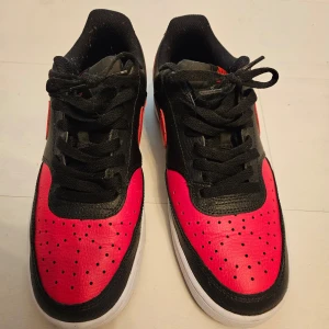 Nike Court Vision i svart och röd. Stl 41 - Snygga Nike sneakers i svart och röd med klassisk design. Skorna har en svart bas med röda detaljer och en vit sula. Perfekta för en sportig look. Storlek 41.