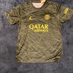 Svart fotbollströja från Jordạn x PSG - Snygg svart fotbollströja från Jordan i samarbete med PSG. Tröjan har ett unikt mönster i gult och korta ärmar. Perfekt för fotbollsfans som vill ha något speciellt. Logotypen för Qatar Airways pryder framsidan.