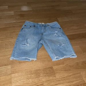 Blå jeansshorts från Levi's - Säljer ett par coola blå jeansshorts från Levi's, modell 511 Slim. Shortsen har en avslappnad look med slitna detaljer och fransiga kanter. Perfekta för en avslappnad stil.