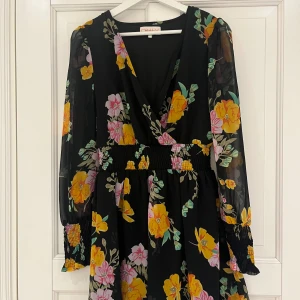 Blommig klänning från only - Snygg blommig klänning från only med v-ringning och långa ärmar. Klänningen har ett färgglatt mönster med gula och rosa blommor på en svart bakgrund. Den har en smockdetalj i midjan och vid ärmsluten som ger en fin passform.