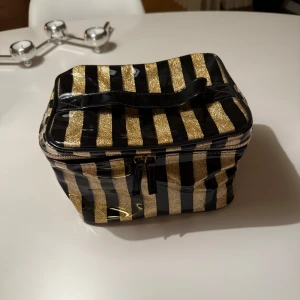 Svart och guld randig väska - Snygg väska med svart och guld randigt mönster. Perfekt för att förvara smink eller andra småsaker. Väskan har en praktisk dragkedja och ett handtag på toppen för enkel transport.