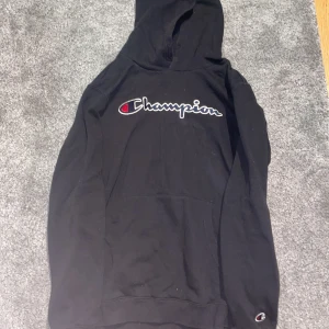 Svart hoodie från Champion - Säljer en svart hoodie från Champion med det klassiska logotyptrycket på bröstet. Hoodien har en känguruficka och är perfekt för en avslappnad stil. Den är tillverkad i mjukt material för extra komfort.