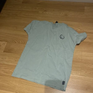 Ljusgrön t-shirt med smiley - Säljer en ljusgrön t-shirt från Great Basics med en svart smiley på bröstet. T-shirten har korta ärmar och en enkel design som passar perfekt för vardagsbruk.