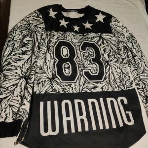 Svart och vit tröja med tryck från ThePower - Säljer en cool svart och vit tröja från ThePower med tryck av siffran 83 och texten 'WARNING'. Tröjan har ett unikt bladmönster och stjärnor på axlarna. Den har långa ärmar och  dragkedjan på sidor för extra stil.