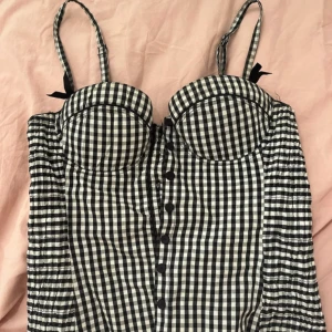 Svartvit rutig korsett från 6ixty8ight - Säljer en snygg svartvit rutig korsett från 6ixty8ight. Den har knappar framtill och justerbara axelband med små rosetter. Perfekt för att ge en stilren look med en touch av vintagekänsla.