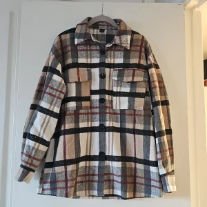 Rutig overshirt från Shein - Säljer en snygg rutig overshirt från Shein i beige, svart och röd. Jackan har långa ärmar och knappar framtill. Perfekt för lager-på-lager-stil och tillverkad i 100% polyester.