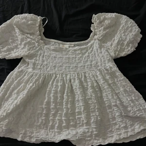 Vit blus från Gina Tricot - Säljer en söt vit blus från Gina Tricot med puffiga korta ärmar och en luftig passform. Tyget har en texturerad yta som ger en fin detalj. Köpt för 300kr