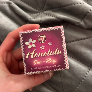 W7 Honolulu Sun-Rays Bronzer - Säljer en W7 Honolulu Sun-Rays bronzer i en söt rosa förpackning med blommönster. Perfekt för att ge ansiktet en solkysst look. Produkten väger 6g och är lätt att applicera för en naturlig glöd.