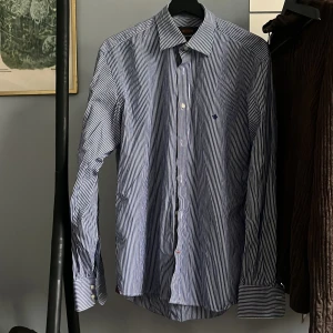 Randig skjorta från Morris - Snygg blå och vit randig skjorta från Morris. Skjortan har långa ärmar och en klassisk krage. Den är broderad med en liten detalj på bröstet och har knappar framtill. Perfekt för en stilren look. Skick 9/10 