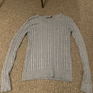 Grå stickad tröja från KappAhl - Säljer en stilren grå stickad tröja från KappAhl. Tröjan har ett klassiskt kabelstickat mönster och långa ärmar. Perfekt för kyligare dagar.