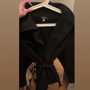 Svart kappa från Boohoo - Snygg svart kappa från Boohoo med knytband i midjan. Perfekt för vår/höst och vinter med en varm tröja under🩷passar M/S