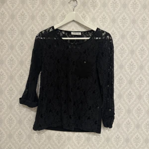 Svart spetsblus från Suiteblanco - Snygg svart spetsblus från Suiteblanco i storlek XS. Blusen har trekvartsärmar och en liten bröstficka med knappdetalj. Perfekt för en elegant look med sin genombrutna spetsdesign.