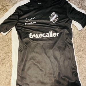 Svart AIK tröja från Nike - Snygg svart fotbollströja från Nike med AIK-logga och Truecaller som sponsor. Tröjan är kortärmad och har vita detaljer längs sidorna. Perfekt för fotbollsfans som vill visa sitt stöd. 