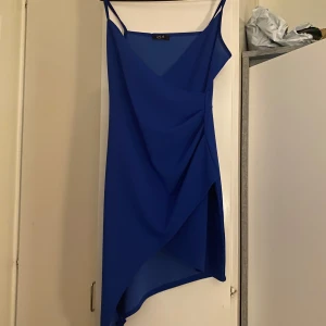 Blå asymmetrisk klänning från ELA - Snygg blå klänning från ELA med asymmetrisk design och smala axelband. Perfekt för en stilren look. Klänningen har en elegant drapering framtill som ger en unik touch.