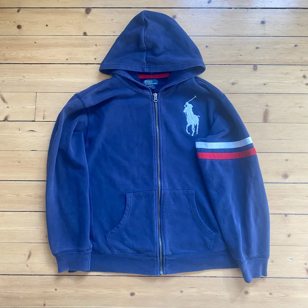 Säljer en snygg blå hoodie från Polo Ralph Lauren. Den har en dragkedja framtill och en stor logga på bröstet. Ärmen har coola ränder i vitt och rött. Perfekt för en avslappnad stil! Storlek L(14-16) sitter som slim S. Tröjor & Koftor.