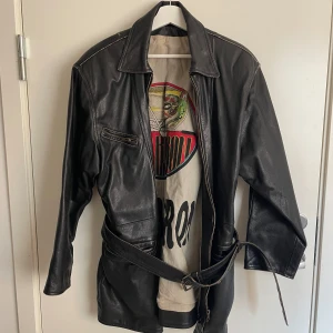 Skinnjacka RED BARON - Pilotjacka i skinn. Den är vintage så märket är oklart men den är onekligen av extremt bra kvalitet. Den är svart/väldigt mörk brun och har en jättesnygg bild på innerfodret samt ett skärp man kan knyta för en mer figursittande passform.  