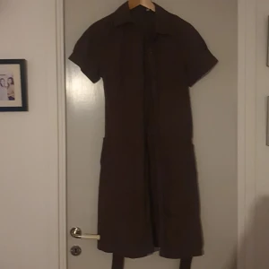 Brun kortärmad klänning - Säljer en brun skjortklänning med korta ärmar och knappar framtill. Klänningen har en klassisk krage och är i ett bekvämt material.