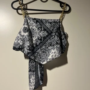 Svartvit mönstrad topp med kedjeband - Snygg svartvit topp med ett coolt paisleymönster. Den har kedjeband som ger en extra touch av stil. Toppen är axelbandslös och har en knytning framtill som gör den perfekt för festliga tillfällen.
