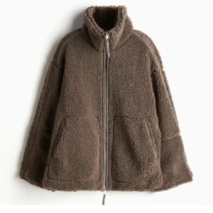Brun teddyjacka med dragkedja - Mysig brun teddyjacka med dragkedja och stora fickor framtill. Perfekt för kyliga dagar med sin fluffiga och varma design. Jackan har en hög krage och en avslappnad passform.
