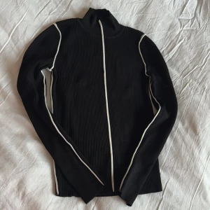 Svart ribbad tröja med vita detaljer - Basic svart ribbad tröja med vita detaljer. Tröjan har en hög krage och långa ärmar, perfekt för en stilren look.