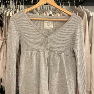 Grå babydoll top - Säljer en stilren grå babydoll kofta med ribbad överdel och knappar framtill. Koftan har långa ärmar och en v-ringad design, perfekt för lager-på-lager outfits. 🌸❄️ hör av er om prisförslag! Passar nog perfekt stl. S (minsta prisförslag 300kr)