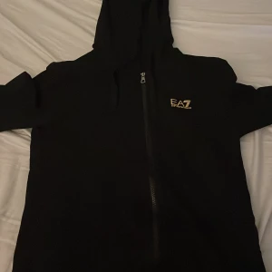 Svart huvtröja från Emporio Armani - Snygg svart huvtröja från Emporio Armani med dragkedja och EA7-logga på bröstet. Perfekt för en stilren och bekväm look.