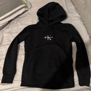 Svart hoodie från Calvin Klein - Snygg svart hoodie från Calvin Klein med logga på bröstet. Den har en klassisk design med huva och dragsko samt en stor magficka. Perfekt för en avslappnad stil.