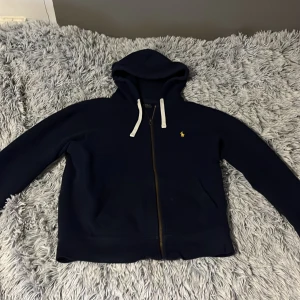 Mörkblå hoodie från Polo Ralph Lauren - Säljer en snygg mörkblå hoodie från Polo Ralph Lauren med dragkedja och huva. Den har en broderad gul logotyp på bröstet och ribbade muddar. Perfekt för en avslappnad stil. (Tröjans storlek är M men passar till S också)