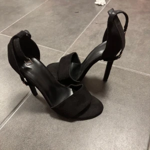 Svarta klackskor med rem - Eleganta svarta klackskor med en smal rem runt ankeln och öppen tå. Perfekta för en kväll ute eller en speciell tillställning. Fint skick, aldrig använda